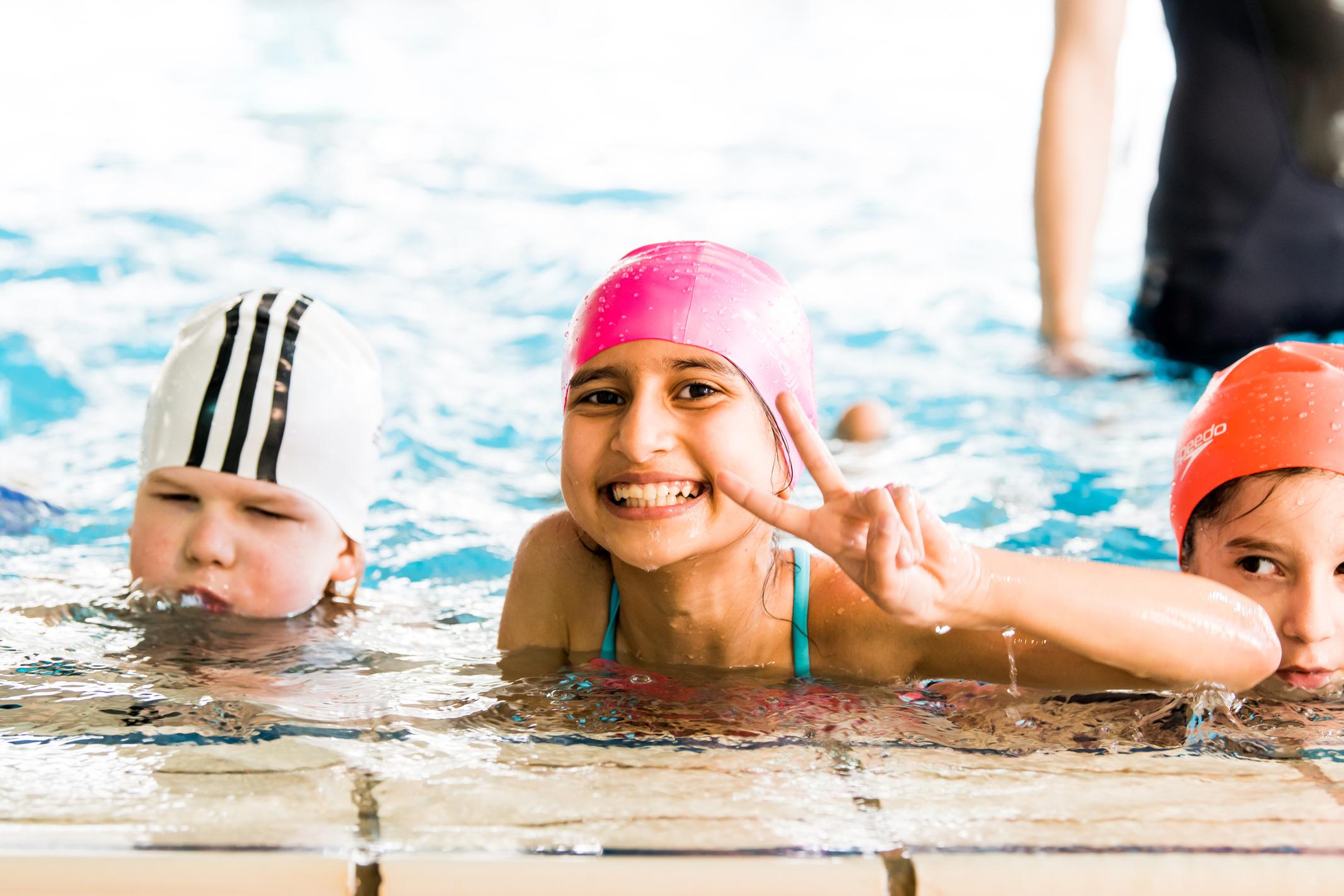 Lächelndes Mädchen mit pinker Badehaube zeigt V-Zeichen im Wasser; zwei weitere Kinder sind im Hintergrund zu sehen.