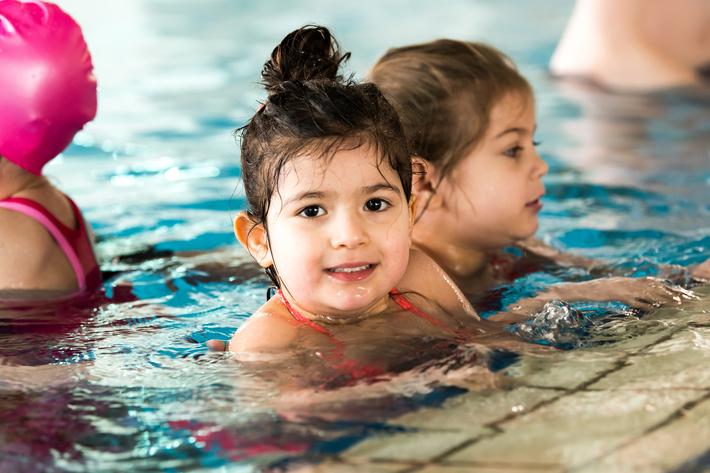 Lächelndes Mädchen mit nassen Haaren im Wasser, im Hintergrund ein weiteres Kind in der Schwimmhalle.